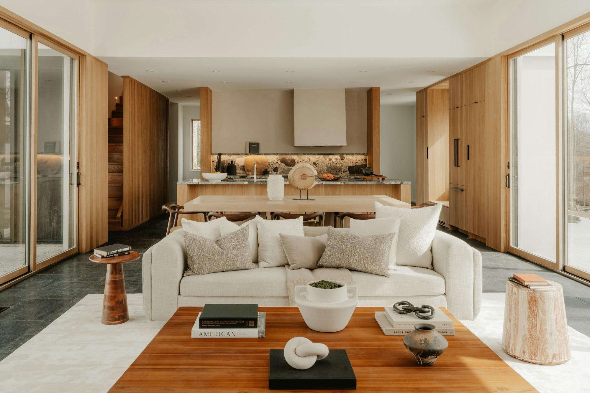 Open-plan living room and kitchen after renovation, with white oak cabinetry, a stone-topped island and full-height glazing to the trees beyond.